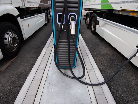 Nahaufnahme einer Elektroladesäule für LKWs zwischen zwei weißen Zugmaschinen. Das Ladekabel ist angeschlossen und die Ladesäule hat eine blaue Beleuchtung.