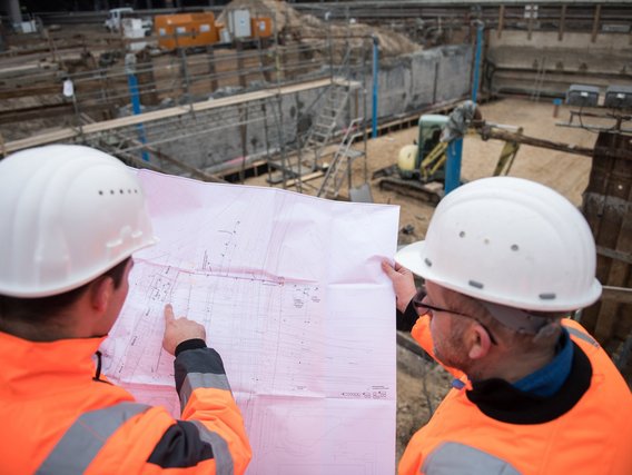 Zwei Bauarbeiter in Sicherheitsausrüstung studieren einen Bauplan, während sie auf eine Baugrube mit Betonwänden blicken.