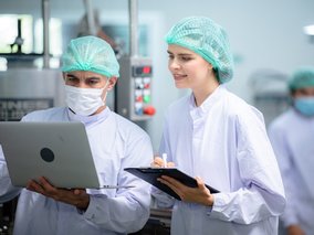 Zwei Personen in weißen Kitteln und Hauben arbeiten mit einem Laptop und einem Klemmbrett in einem sterilen Raum.