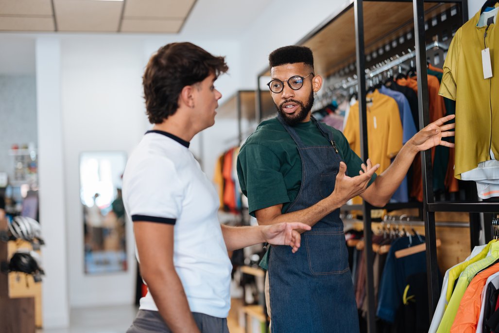 Ein Verkäufer in einem Sportbekleidungsgeschäft zeigt einem Kunden verschiedene Kleidungsstücke. Der Verkäufer trägt eine grüne Schürze und gestikuliert, während er dem Kunden die Auswahl erklärt. Im Hintergrund sind Regale mit bunten Sportshirts zu sehen.