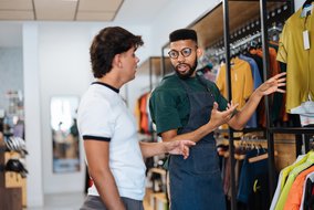 Ein Verkäufer in einem Sportbekleidungsgeschäft zeigt einem Kunden verschiedene Kleidungsstücke. Der Verkäufer trägt eine grüne Schürze und gestikuliert, während er dem Kunden die Auswahl erklärt. Im Hintergrund sind Regale mit bunten Sportshirts zu sehen.