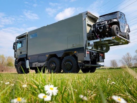 Ein grauer Lkw steht auf einer Wiese und transportiert auf seiner Ladefläche ein schwarzes Auto. Auf dem Boden wachsen kleine weiße Blumen.