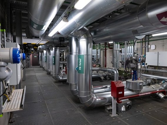 Ein hochmoderner Maschinenraum mit silbernen Rohrleitungen, Ventilen und technischen Anlagen unter Neonbeleuchtung. Die industrielle Anlage mit Metallgittern am Boden und grünen Schildern dient der Gebäude- oder Anlagentechnik.