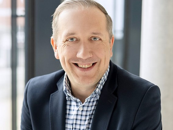 Tobias Mielke im dunklen Anzug und kariertem Hemd vor hellem Büro-Hintergrund mit Fenstern.