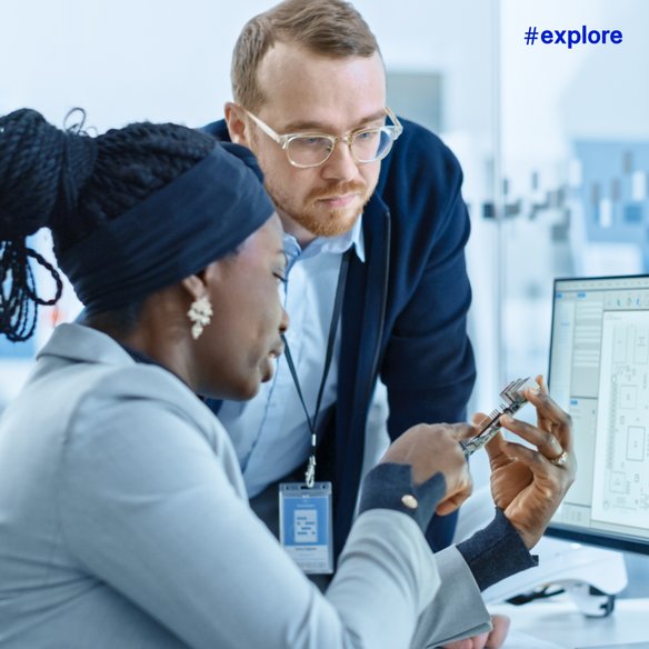 Zwei Personen in einem modernen Büro diskutieren über technische Zeichnungen auf einem Computerbildschirm. Eine Person hält ein elektronisches Bauteil in der Hand. Der Hashtag "#explore" ist oben rechts im Bild zu sehen.