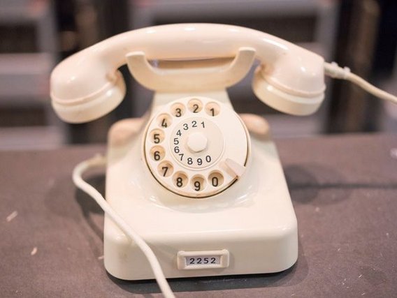 Ein cremefarbenes, klassisches Wählscheibentelefon mit einem runden Ziffernblatt und einem angeschlossenen Hörer. Die Nummern sind in einem Kreis angeordnet, und ein Kabel verbindet das Gerät mit dem Telefonnetz.