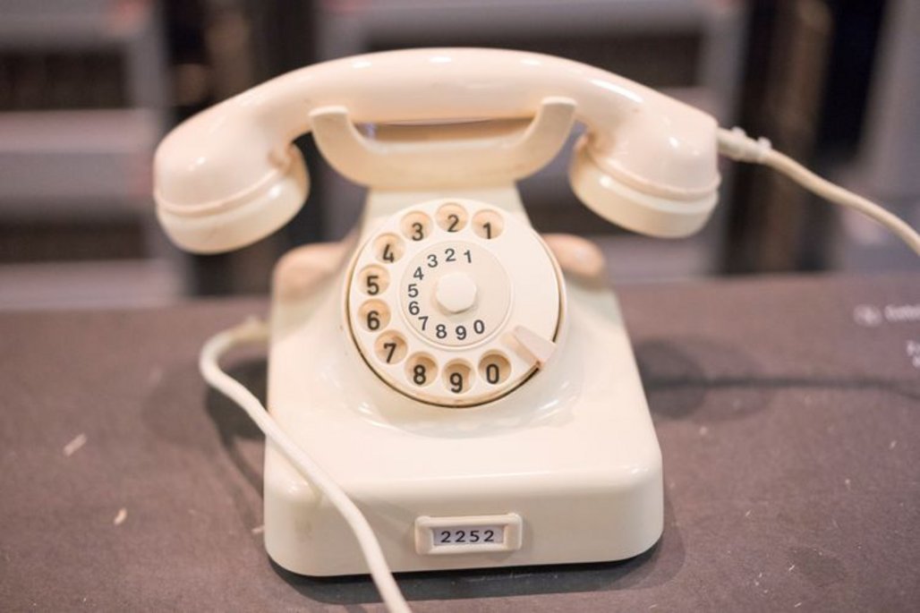 Ein cremefarbenes, klassisches Wählscheibentelefon mit einem runden Ziffernblatt und einem angeschlossenen Hörer. Die Nummern sind in einem Kreis angeordnet, und ein Kabel verbindet das Gerät mit dem Telefonnetz.