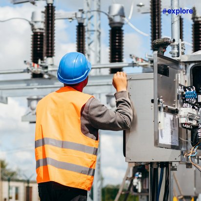 Eine Person in einer orangefarbenen Warnweste und einem blauen Schutzhelm steht vor einem Schaltschrank in einem Umspannwerk. Sie arbeitet an elektrischen Komponenten, die von Kabeln und Schaltern umgeben sind. Im Hintergrund sind große Isolatoren und Stromleitungen zu sehen. Der Himmel ist leicht bewölkt.