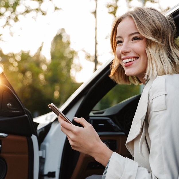 Lächelnde junge Frau sitzt mit Smartphone in der Hand in der offenen Tür eines Autos