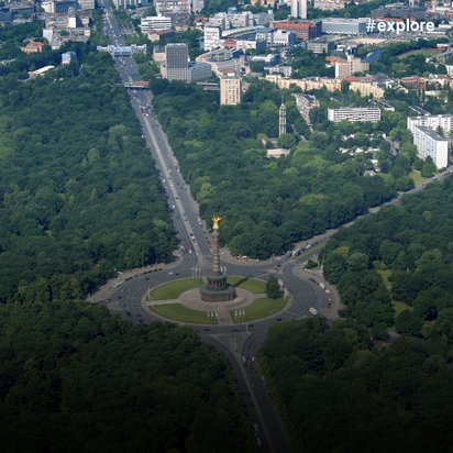 Eine Luftaufnahme zeigt eine deutsche Stadt mit einem kreisförmigen Platz und goldener Säule im Zentrum, umgeben von grünen Parks. Die städtische Infrastruktur ist harmonisch in die Landschaft integriert.