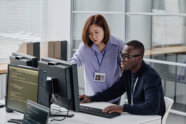 Zwei IT-Fachkräfte arbeiten gemeinsam an Code auf mehreren Monitoren in einem modernen Büro
