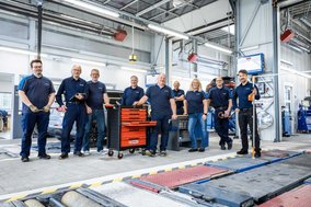 TÜV NORD Werkstatt-Team in Arbeitskleidung steht gemeinsam in der Prüfhalle, umgeben von Werkstattausrüstung.