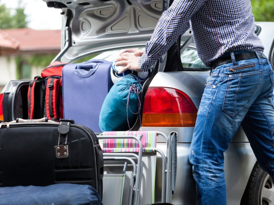 Mann in kariertem Hemd und Jeans packt bunte Koffer und Gepäckstücke in den geöffneten Kofferraum eines silbernen Autos.