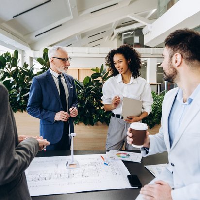 Gruppe von Geschäftsleuten im Gespräch vor Bauplänen in einem modernen Bürogebäude.