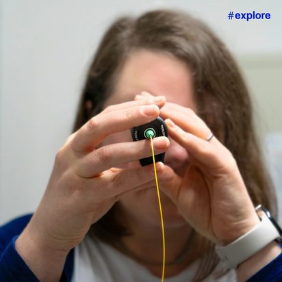 Eine Person hält ein gelbes Kabel und betrachtet es durch ein Gerät. Die Person trägt ein weißes Armband und hat braunes Haar. Oben rechts im Bild ist der Hashtag "#explore" in blauer Schrift zu sehen.