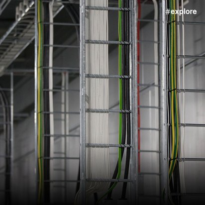 Mehrere silberne Aluminiumleitern stehen in einer Reihe in einem Industriegebäude mit weißen Wänden und sichtbaren Rohrleitungen. Die Leitern haben verschiedene Farben an den Rahmen - grün, rot und andere Akzentfarben.