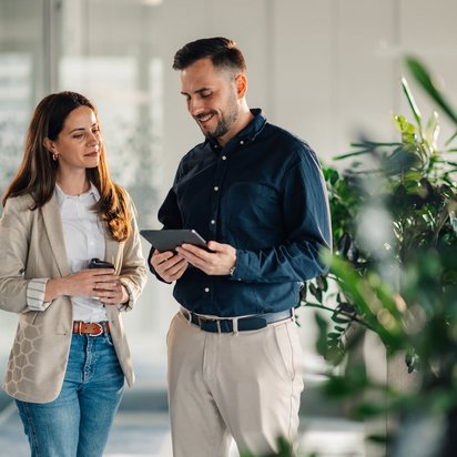 Zwei Kolleg:innen unterhalten sich freundlich im Büroflur mit Tablet und Kaffeebecher