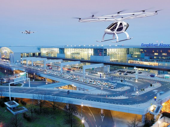 Luftaufnahme des modernen Flughafens Frankfurt mit seiner charakteristischen Glasarchitektur, über dem ein weißes Flugtaxi schwebt.