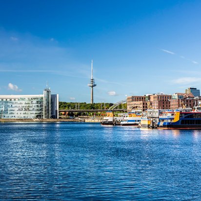 Blick auf den Hafen von Kiel mit modernen Gebäuden und einem Fernsehturm im Hintergrund