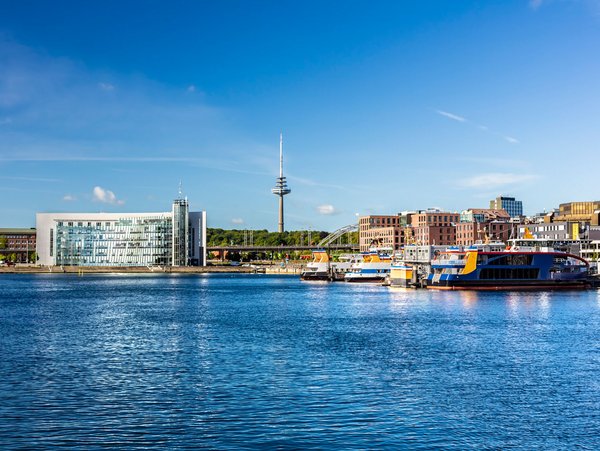 Blick auf den Hafen von Kiel mit modernen Gebäuden und einem Fernsehturm im Hintergrund