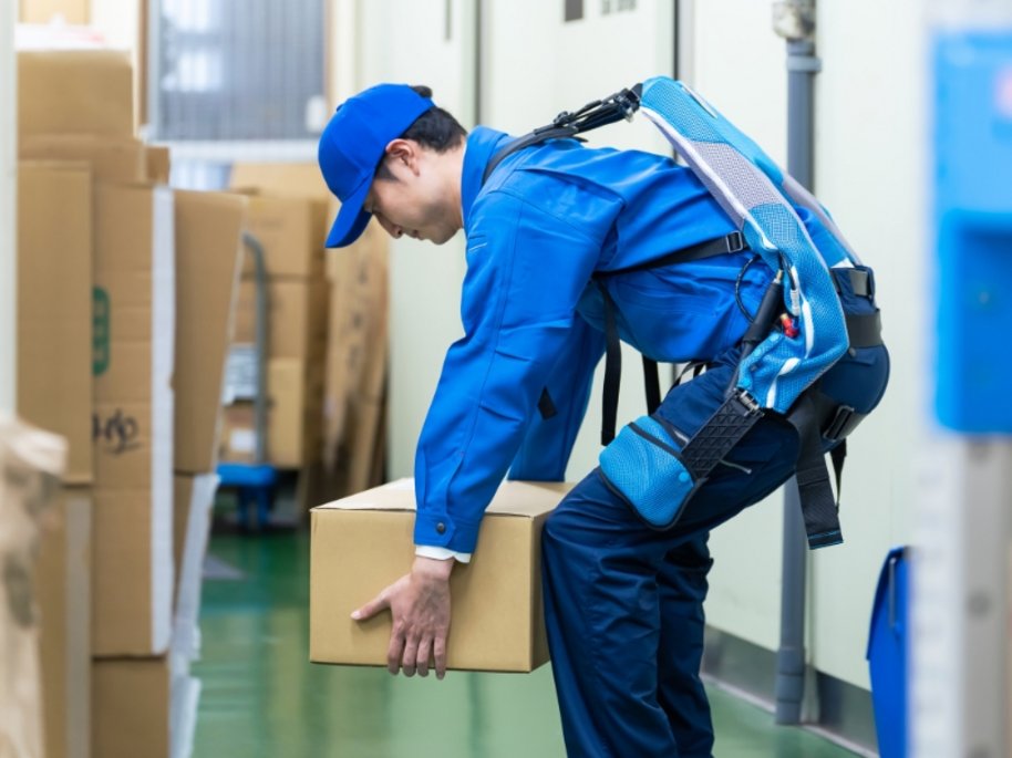 Ein Arbeiter in blauer Uniform und Mütze hebt eine Kartonschachtel in einem Lagerraum mit Regalen und Kartons.
