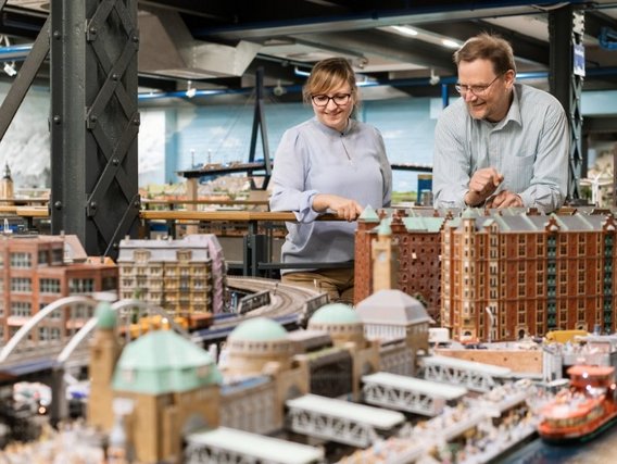 Dr. Hans Vallée und Katrin Leicht mit Brillen betrachten lächelnd eine detaillierte Miniaturstadt mit Gebäuden, Eisenbahnen und einem Hafen in einem Ausstellungsraum.