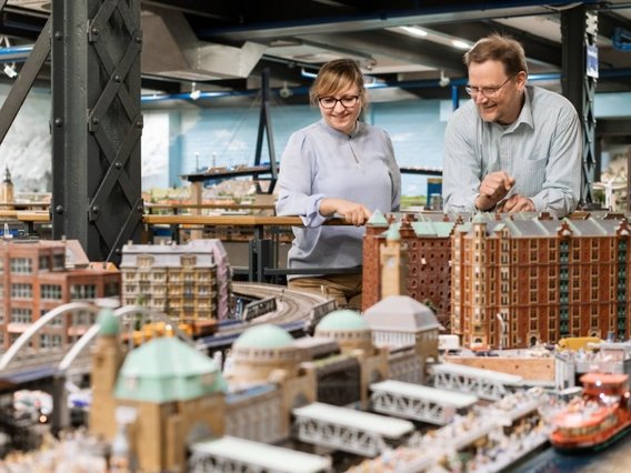 Dr. Hans Vallée und Katrin Leicht mit Brillen betrachten lächelnd eine detaillierte Miniaturstadt mit Gebäuden, Eisenbahnen und einem Hafen in einem Ausstellungsraum.