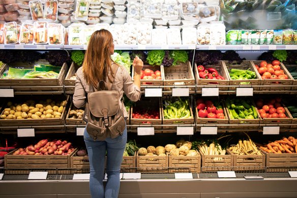 Frau mit Rucksack steht vor einem Gemüse-Regal im Supermarkt und wählt frisches Gemüse aus