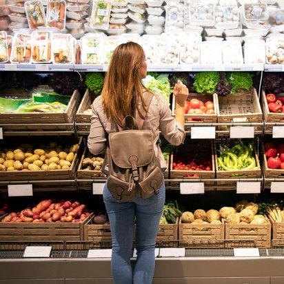 Frau mit Rucksack steht vor einem Gemüse-Regal im Supermarkt und wählt frisches Gemüse aus