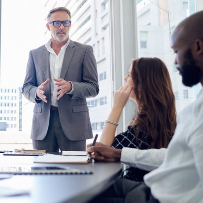Präsentation eines Projektleiters im Business-Meeting. Mann im Anzug spricht vor einer Gruppe von Personen in einem modernen Konferenzraum.