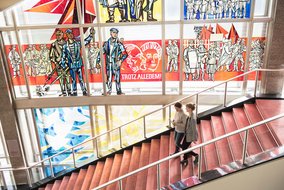 Eine Treppe mit rotem Belag und Metallgeländer führt an einer großen Glaswand mit bunten politischen Motiven und Darstellungen vorbei. Unten auf der Treppe sind zwei Personen zu sehen.