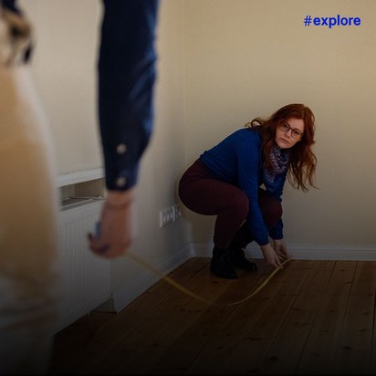 Zwei Personen vermessen einen Raum mit einem Maßband auf einem Holzboden. Eine Frau in blauem Pullover, burgunderroten Hosen und Brille hockt an der Wand, während eine andere Person im Vordergrund teilweise sichtbar ist. Der Raum hat cremefarbene Wände und weiße Fußleisten.