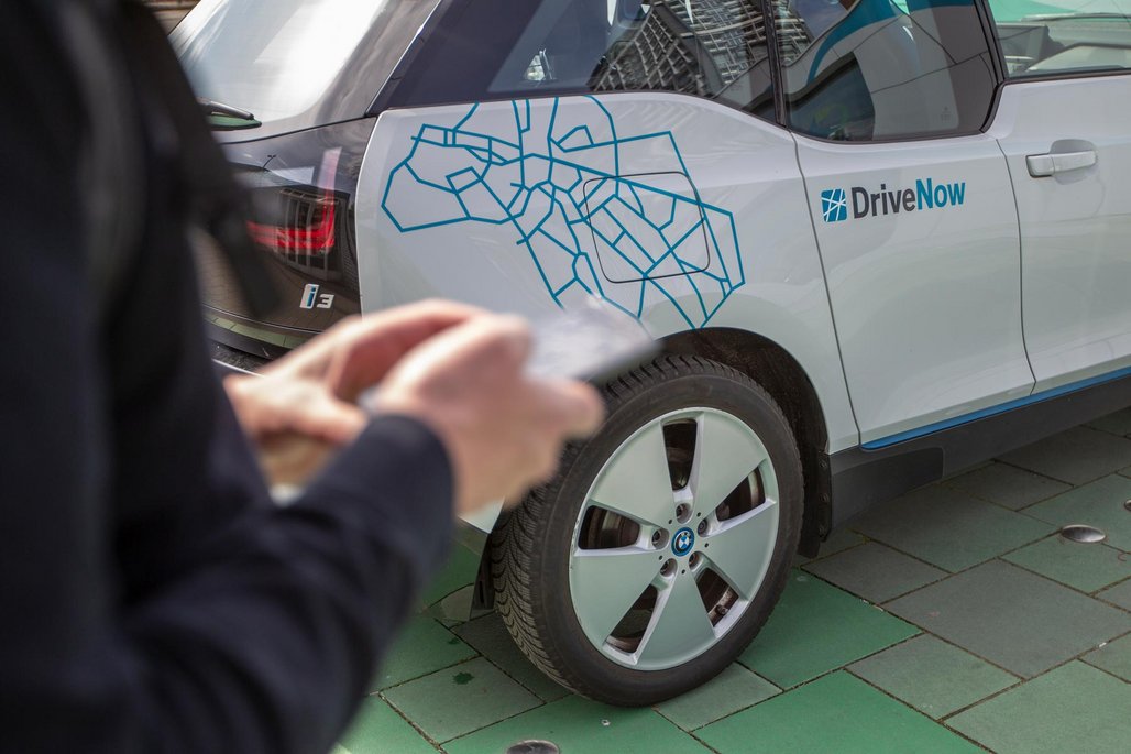 Person entsperrt ein weißes DriveNow Carsharing-Fahrzeug mit dem Smartphone auf einem Gehweg.