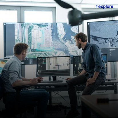 Zwei Männer in einem Kontrollraum mit mehreren Bildschirmen und Karten. Sie analysieren Luftaufnahmen und Geodaten, während im Vordergrund Teile einer Drohne oder Kameraausrüstung zu sehen sind.