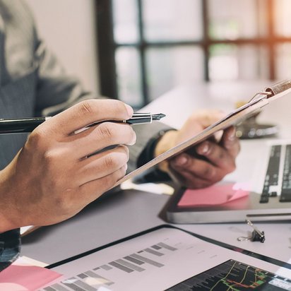 Person prüft ein Dokument auf einem Klemmbrett mit Stift in der Hand, Laptop und Diagramme auf dem Schreibtisch