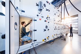 Zwei Techniker arbeiten an einer großen, weißen Lüftungsanlage; einer kniet im Inneren einer Wartungsklappe, während der andere außen kontrolliert.
