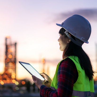 Eine Person in Schutzkleidung steht in einem Industrieviertel und schaut auf ihr Tablet.