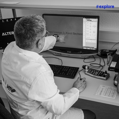 Eine Person in einem Laborkittel sitzt an einem Tisch und zeigt auf einen Bildschirm, während sie ein Mikroskop bedient. Auf dem Tisch befinden sich eine Tastatur, eine Maus und ein Steuergerät. Der Raum ist mit wissenschaftlicher Ausrüstung ausgestattet.