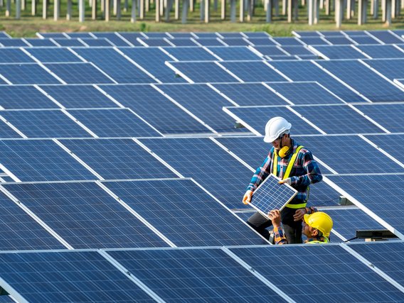 Zwei Arbeiter mit Schutzhelmen installieren Solarmodule auf einem großen Solarpark, einer hält ein Paneel während der andere bei der Montage hilft.