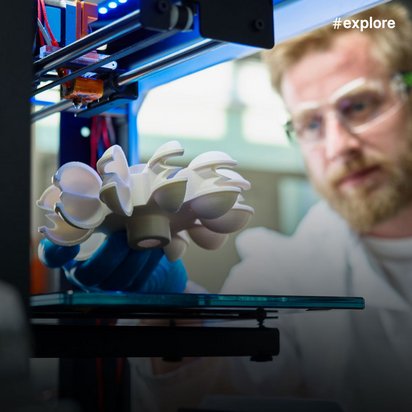 Ein Forscher mit Schutzbrille beobachtet konzentriert einen 3D-Drucker, der gerade ein komplexes weißes Objekt mit organischen, blütenartigen Formen druckt. Das moderne Labor im Hintergrund unterstreicht die fortschrittliche Fertigungstechnologie und präzise Materialforschung.