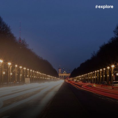 Nächtliche Langzeitbelichtung einer von Bäumen gesäumten Allee mit leuchtenden Lichtspuren von Autos, die zum Brandenburger Tor in Berlin führt. Der tiefblaue Himmel und die warmen Straßenlaternen schaffen eine dramatische Atmosphäre mit dem Hashtag "#explore" in der oberen rechten Ecke