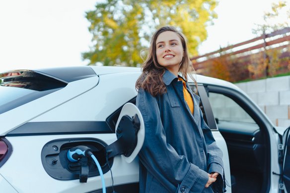 Frau steht neben einem weißen Elektroauto, das an einer Ladesäule angeschlossen ist.