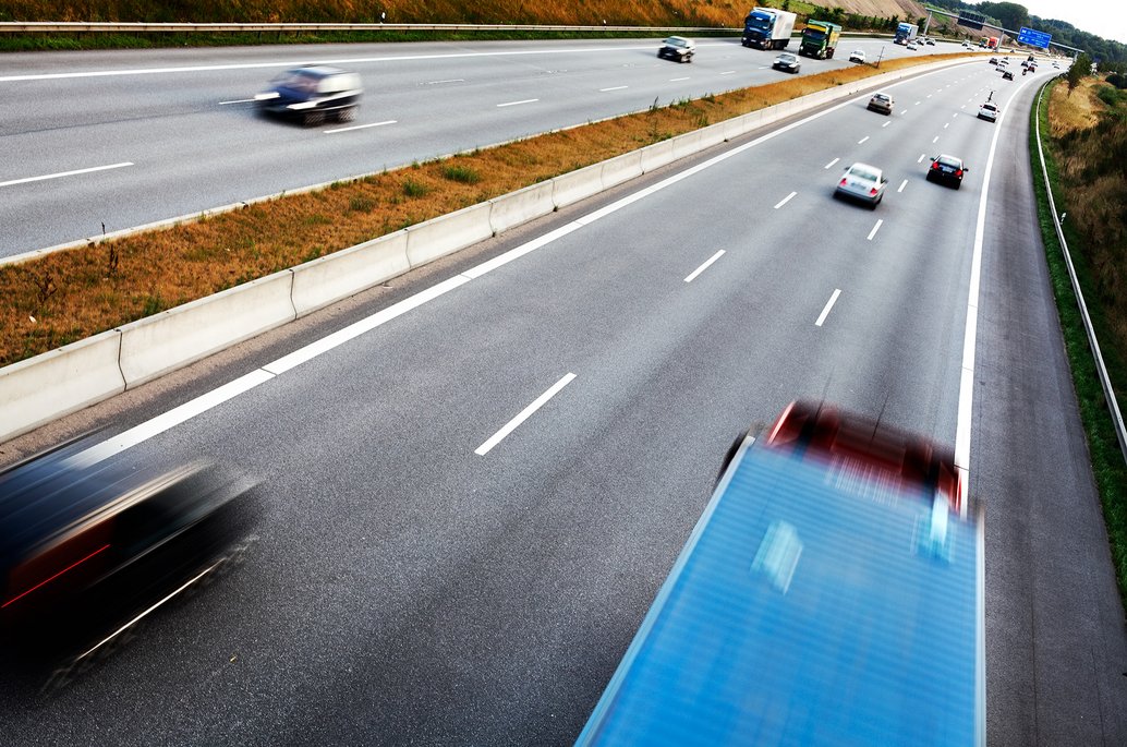 Luftaufnahme einer mehrspurigen Autobahn mit schnell fahrenden Fahrzeugen, die durch Bewegungsunschärfe dargestellt sind. Ein blauer und ein schwarzer PKW sowie LKWs sind auf den verschiedenen Fahrspuren zu sehen.
