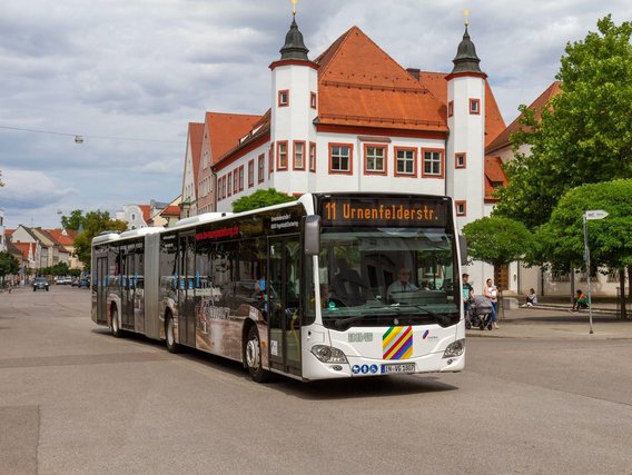 Ein weißer Linienbus der Linie 11 mit Ziel "Urnenfelderstr." steht vor einem historischen Gebäude mit weißer Fassade und roten Dachziegeln. Das Gebäude hat zwei runde Türme und zeigt typische süddeutsche Architektur.