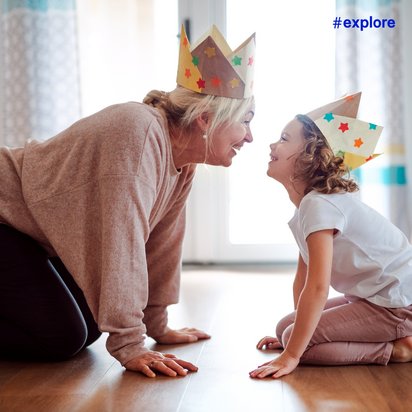 Eine ältere Person und ein Kind tragen selbstgebastelte Papierkronen mit bunten Sternen und knien sich im Wohnzimmer gegenüber. Sie sind auf einem Holzboden und die Umgebung ist hell und freundlich mit Vorhängen und einem Stativ im Hintergrund.