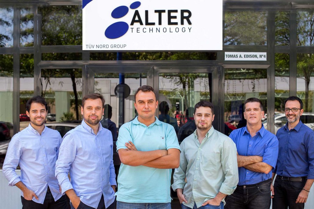 Sechs Männer in Business-Casual-Kleidung stehen vor dem Firmengebäude von Alter Technology. Sie tragen verschiedene Hemden in Blau- und Grüntönen und lächeln in die Kamera.