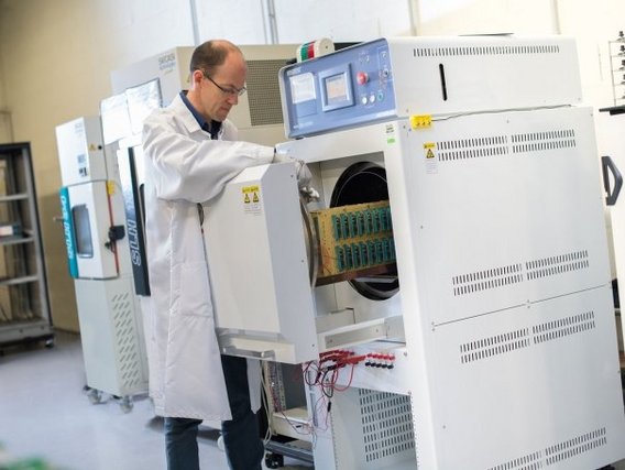 Ein Mann im weißen Laborkittel bedient ein großes Laborgerät mit blauem Display und offener Klappe. Im Inneren des Geräts sind grüne Elektronikkomponenten sichtbar.
