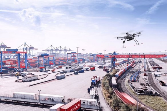 Eine Drohne fliegt über einen großen Containerhafen mit zahlreichen Containern, Kränen und Güterzügen. Die Luftaufnahme zeigt die weitläufige Logistikinfrastruktur unter blauem Himmel.