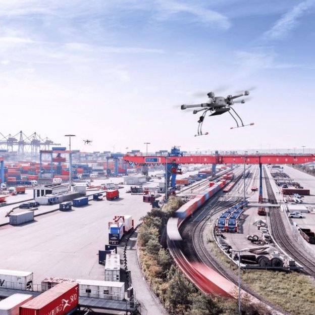 Eine Drohne fliegt über einen großen Containerhafen mit zahlreichen Containern, Kränen und Güterzügen. Die Luftaufnahme zeigt die weitläufige Logistikinfrastruktur unter blauem Himmel.