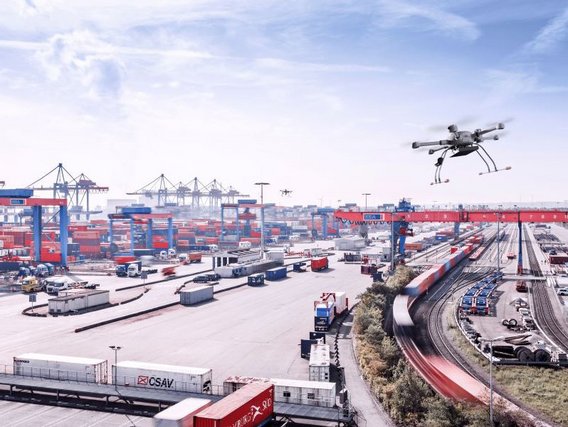 Eine Drohne fliegt über einen großen Containerhafen mit zahlreichen Containern, Kränen und Güterzügen. Die Luftaufnahme zeigt die weitläufige Logistikinfrastruktur unter blauem Himmel.